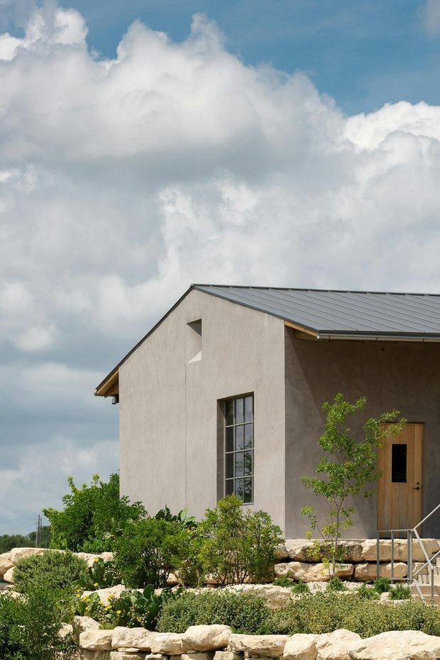Exterior view of a compact rendered house with a simple gabled metal roof, tall narrow divided window, timber entry door, and a landscaped terraced garden with stone retaining walls. The building has a restrained contemporary form and sits beneath a dramatic cloudy sky.