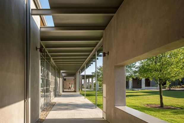 A long, covered architectural walkway between rendered walls, with exposed metal roof beams and narrow skylight openings. Floor-to-ceiling glazed panels line one side, while a large rectangular opening frames a landscaped lawn and trees. The restrained palette and strong linear perspective create a contemporary institutional or commercial character.