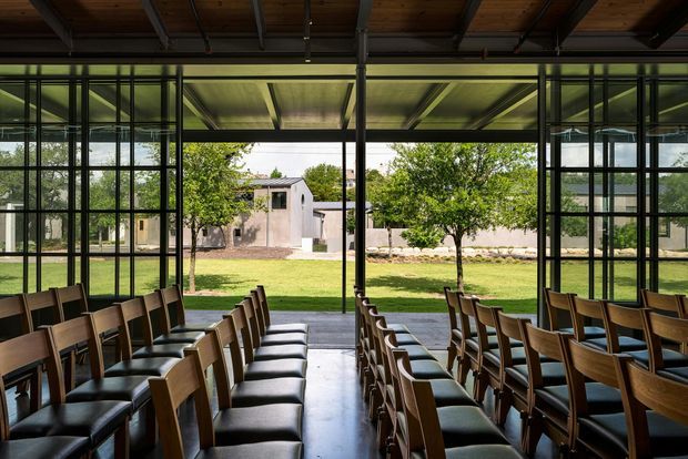 A large, formal interior with rows of timber-framed chairs upholstered in dark leather, arranged facing an unseen focal point. Floor-to-ceiling black metal-framed glazing opens views to a landscaped courtyard and a low masonry building beyond. The space has an exposed timber ceiling with dark structural beams and a polished concrete floor, creating a contemporary, industrial-inspired event or gathering venue.