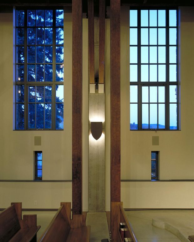 Interior view of a tall, dramatic worship or assembly space with paired multi-pane windows, exposed dark timber structural posts, pale plaster walls, polished concrete or stone flooring, and timber pews in the foreground. A narrow illuminated vertical feature with a shield-like sculptural element forms a central focal point. The high ceiling and monumental vertical composition create a solemn, contemplative atmosphere.