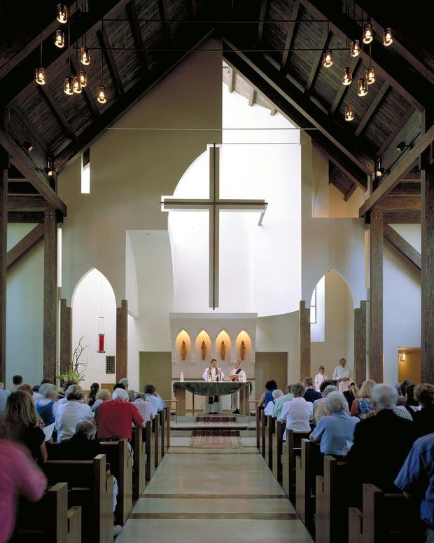 Interior view down the central aisle of a church or chapel during a service. The space features a dramatic double-height vaulted timber roof with exposed dark timber beams and rafters, suspended pendant lights, white plastered walls, tall arched openings, a large central cross, altar, and rows of pews occupied by congregants.