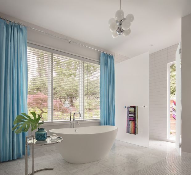 Bright, spacious bathroom featuring a freestanding oval bathtub positioned in front of a large window with white blinds and vivid blue curtains. The room has pale marble-look flooring, white horizontal wall tiling, a wall-mounted towel rack with a colourful textile, and a chrome-and-glass side table with a potted plant. A sculptural cluster pendant light hangs from the ceiling, and an open doorway leads to an adjacent area.