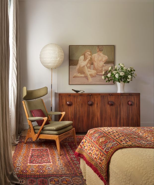 A warmly styled bedroom corner with a mid-century lounge chair and ottoman beside a tall textured globe floor lamp. A dark timber sideboard with round drawer pulls sits against a pale wall beneath framed artwork and a vase of flowers. Layered patterned rugs and a richly patterned textile draped over the bed add strong colour and texture, while full-height curtains frame the window.