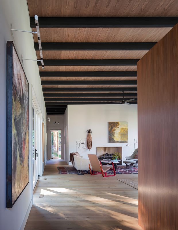 A bright, spacious contemporary living room viewed from a hallway or entry. The room features a warm timber floor, a dramatic ceiling with exposed dark beams and timber infill panels, white walls, large artwork, a fireplace, patterned rug, and mid-century-inspired lounge chairs. Full-height timber wall panelling or joinery frames the right side, while glazed doors and windows line the left.