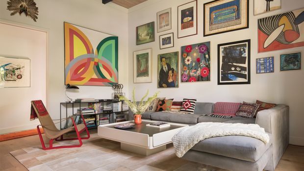 A bright, eclectic living room featuring a large grey sectional sofa, white geometric coffee table, red-framed lounge chair, open shelving filled with books, and a densely arranged gallery wall of colourful artwork. The room has pale walls, light wood flooring, a timber ceiling detail, and a relaxed, art-focused character.