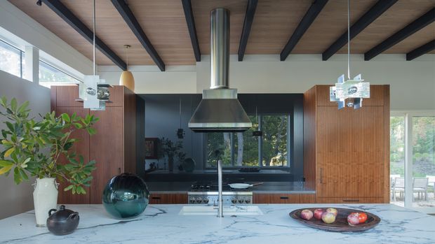 A spacious, refined contemporary kitchen with a large white veined stone island, integrated cooktop and sink, warm vertical timber cabinetry, dark blue-black lower cabinetry and backsplash, and a dramatic suspended stainless-steel rangehood. The room has a timber-lined ceiling with exposed dark beams, large windows, and sculptural pendant lights, creating a warm, high-end modern interior.