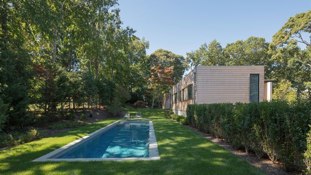 Exterior view of a contemporary single-storey residence set within a lush landscaped garden. A long rectangular swimming pool occupies the foreground beside a manicured lawn, while the house features horizontal timber cladding, large vertical glazing, and a simple low-profile form partially screened by hedges and mature trees.