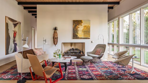 A warm, eclectic living room with floor-to-ceiling glazing along one side, a prominent central fireplace, exposed dark ceiling beams, and a richly patterned layered rug. The room contains mid-century-inspired lounge chairs, a sofa, sculptural lighting, artwork, and natural timber details, creating a relaxed yet curated atmosphere.