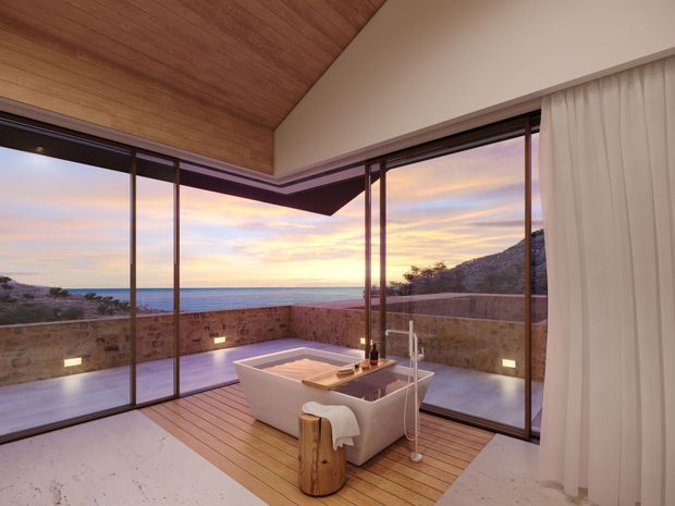 Luxurious freestanding bathtub positioned in a glazed corner room overlooking a coastal seascape at sunset. The space features extensive floor-to-ceiling glazing with dark slim frames, warm timber ceiling and decking, pale stone-like flooring, a low natural stone terrace wall, and sheer white curtains. The open corner creates an airy, serene indoor-outdoor bathing environment.