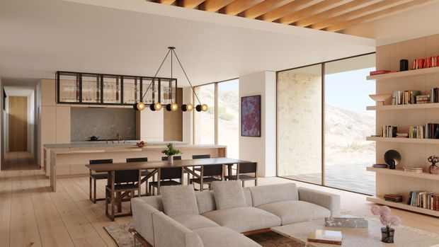 Open-plan contemporary interior combining a kitchen, dining area, and living room. The space features a pale timber floor, warm neutral finishes, a large kitchen island, dark timber dining furniture, a light sectional sofa, extensive floor-to-ceiling glazing, built-in shelving, and exposed timber ceiling rafters. A mountain landscape is visible through the glazing.