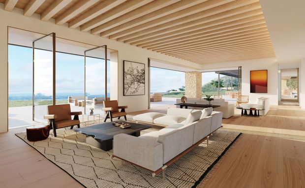 A bright, spacious contemporary living room with an open-plan layout, expansive floor-to-ceiling glazing, and views toward the coastline. The room features a large light upholstered sectional sofa, dark rectangular coffee table, patterned area rug, sculptural lounge chairs, and a minimalist console. Warm timber flooring and a ceiling of closely spaced exposed timber battens create a strong natural architectural feature, while broad openings connect the living area to an outdoor terrace.