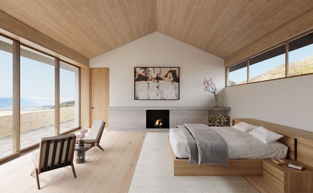 A serene coastal bedroom with a low timber platform bed, soft grey bedding, extensive floor-to-ceiling glazing, and a minimalist fireplace set into a pale stone or concrete surround. Warm timber lines the pitched ceiling and flooring, while ocean views and a rocky landscape are visible outside. A built-in timber bedside ledge and firewood storage reinforce the understated natural design.