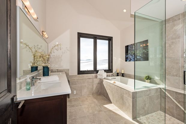 Bright contemporary bathroom with a freestanding-style built-in bathtub set into a tiled surround, a frameless glass shower enclosure, double vanity with dark wood cabinetry, large wall mirror, and dark-framed window overlooking a coastal scene. Neutral beige and white finishes create a clean, refined feel.