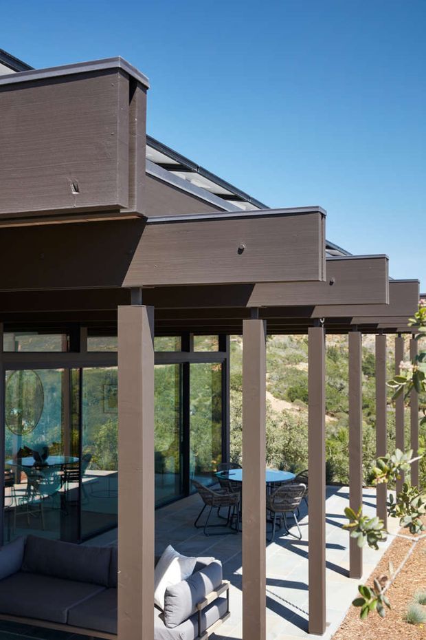 Exterior view of a contemporary multi-level building with projecting dark brown cladding volumes, deep roof overhangs, and a long covered outdoor terrace. Slim square columns frame extensive dark-framed glazing, with outdoor dining and lounge furniture visible beneath the canopy. The structure is set against a dry, landscaped hillside under a clear blue sky.