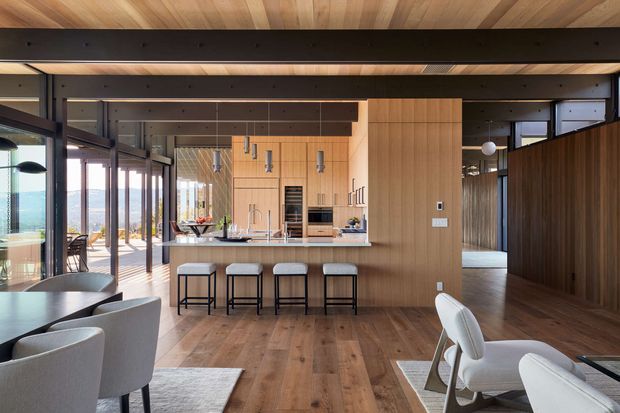 Open-plan contemporary kitchen and dining/living area with extensive glazing, warm timber wall and ceiling finishes, a central island with four stools, integrated appliances, and dark structural beams. The space opens toward a bright exterior view and includes a dining table and upholstered lounge chairs.