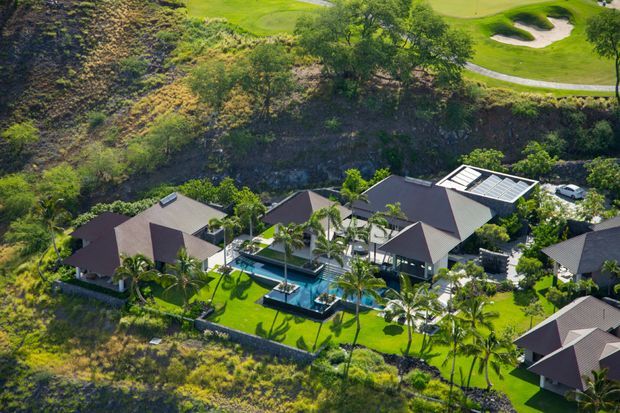 Aerial view of a large resort-style residential property set within a lush, landscaped site. Multiple dark pitched-roof buildings surround a central swimming pool and bright green lawns, with numerous palm trees and a steep vegetated hillside behind. A golf-course-like landscape and winding paved path are visible in the distance.