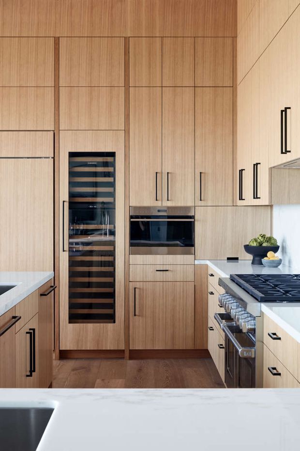 A compact, highly finished contemporary kitchen with floor-to-ceiling light timber cabinetry, integrated appliances, a dedicated glass-front wine cooler, dark linear handles, and pale stone benchtops. A large gas cooktop and stainless-steel range occupy the right-hand run, while a sink is visible in the foreground island. Warm timber flooring and cabinetry create a refined, cohesive look.