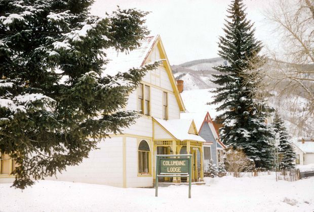 Snow-covered exterior of Columbine Lodge, a steep-gabled timber-clad building surrounded by evergreen trees in a mountainous winter setting. The facade features pale weatherboard-style cladding, contrasting yellow trim, arched windows, and a projecting entry porch with a lodge sign.