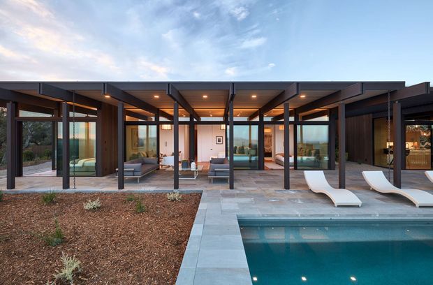 A wide exterior view of a contemporary single-storey residence opening onto a covered pool terrace. The home has a long, low flat roof with extensive floor-to-ceiling glazing, dark structural posts and frames, warm timber-toned soffits, and a paved outdoor entertaining area. A rectangular swimming pool with sculptural loungers occupies the foreground, while landscaped garden beds sit beside the building.