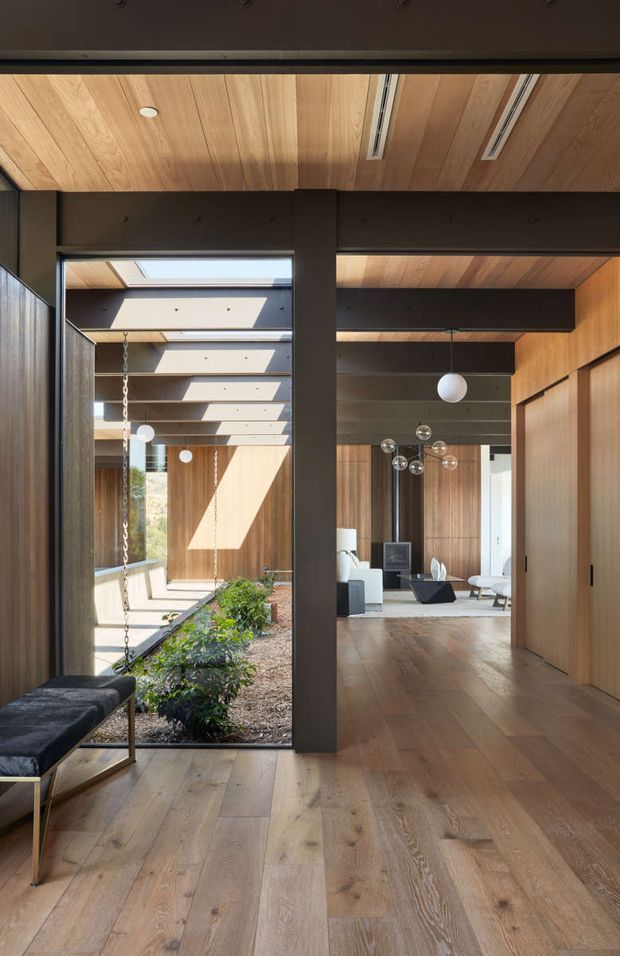 A refined contemporary interior hallway or living space with extensive warm timber lining, wide timber flooring, dark structural beams and framing, and large glazed openings overlooking a planted courtyard. A skylit passage leads toward a furnished living area with sculptural pendant lighting, while a built-in timber wall conceals storage or doors. The space has strong indoor-outdoor connection, abundant natural light, and exposed structural detailing.