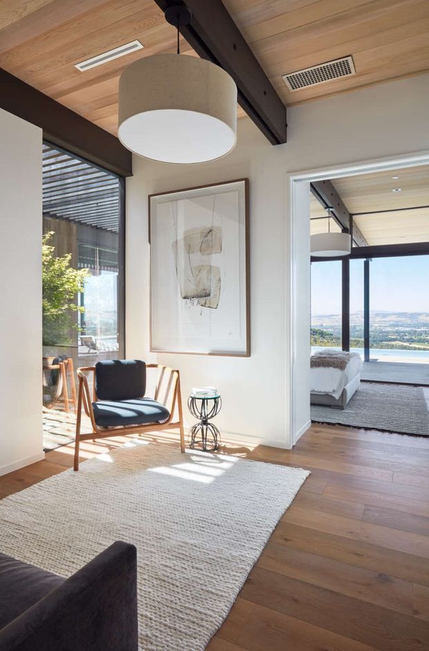 Sunlit contemporary interior showing a relaxed sitting area with a timber-framed lounge chair, small metal side table, large pale textured rug, framed artwork, and part of a dark sofa. The space opens toward a bedroom with expansive glazing and a scenic landscape view. Exposed dark ceiling beam, timber ceiling lining, pale walls, and timber flooring create a warm modern character.