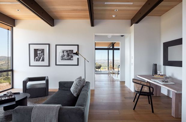 A refined contemporary living room with a dark charcoal sectional sofa, matching armchair, framed black-and-white artwork, and a slim desk or console along the right wall. The room features warm timber ceiling boards with exposed dark beams, pale plaster walls, dark timber flooring, and large windows overlooking a distant landscape. An open doorway leads to a bright dining area with extensive glazing and outdoor views.