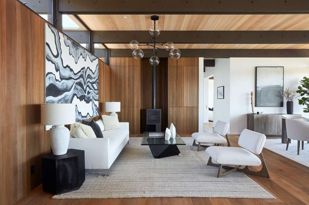 A refined open-plan living room with warm vertical timber wall panelling and a timber-lined ceiling crossed by dark exposed beams. The room features a white sofa, sculptural lounge chairs, a black geometric coffee table, neutral rug, stone-like lamps, and a large abstract monochrome artwork. A dining area is visible beyond the living space, with white walls, timber flooring, and contemporary furnishings. Full-height glazing and strong horizontal and vertical timber lines create a bright, sophisticated interior.