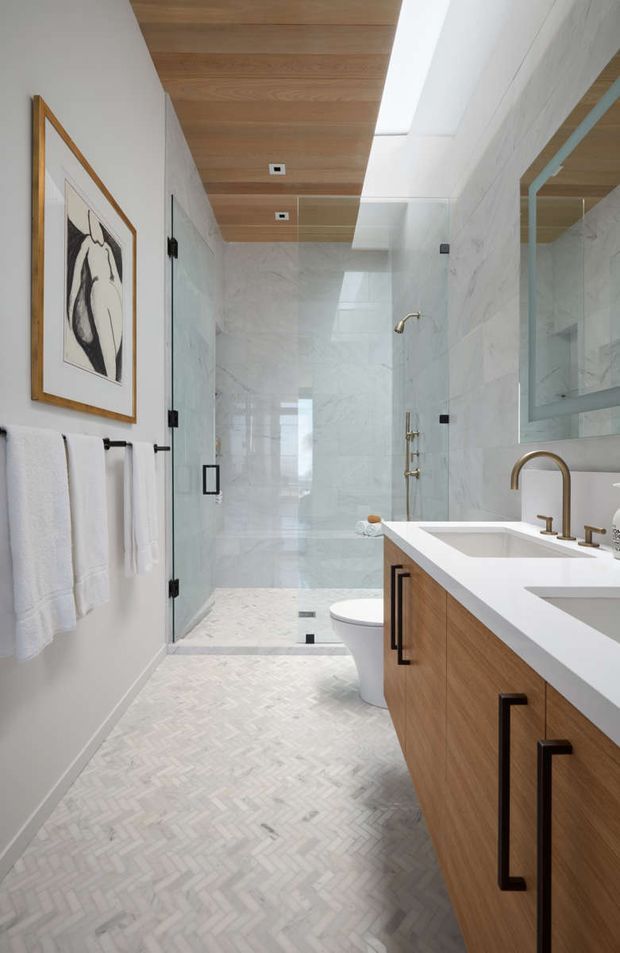 Bright contemporary bathroom featuring a large glass-enclosed walk-in shower, white marble-look wall slabs, a white herringbone floor, and a timber double vanity with a white benchtop and brass fixtures. A warm timber ceiling, skylight, frameless glass panels, towel rails, and framed artwork complete the refined scheme.