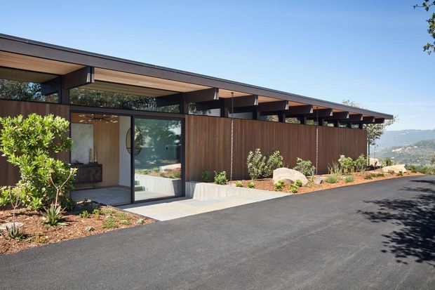 Single-storey modern residence viewed from the driveway, featuring a long, low-pitched roof with deep overhangs, exposed timber roof framing, extensive glazing, and vertical timber cladding. A landscaped garden and paved entry sit beside the dark asphalt driveway, with rolling hills visible in the distance.