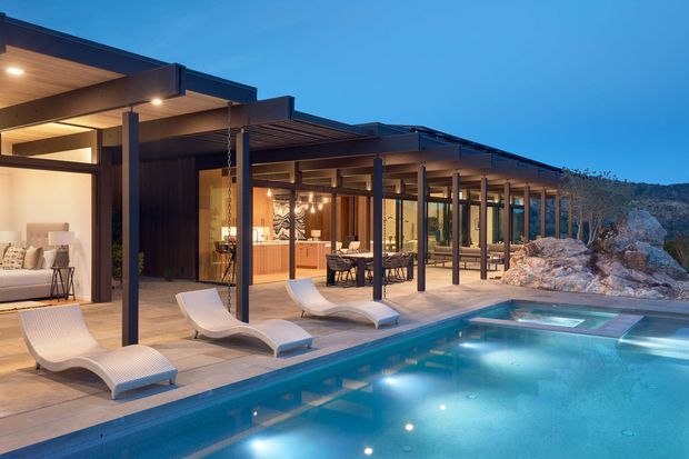 A twilight exterior view of a low-slung contemporary residence arranged around a swimming pool. Broad covered terraces with dark slender steel or aluminium posts, deep flat roof planes, extensive floor-to-ceiling glazing, and warm timber soffits connect the indoor living areas to the pool deck. A warmly lit kitchen and dining area are visible through the glazing, while lounge furniture and sculptural poolside loungers occupy the terrace. Rocky natural terrain borders the property.