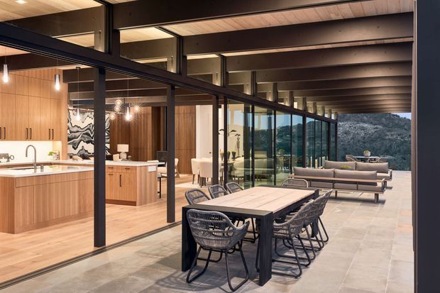 A refined open-plan residential interior with a kitchen, dining area and living room arranged beneath exposed dark structural beams. Floor-to-ceiling black-framed glazing opens toward a rocky natural landscape. The kitchen features warm timber cabinetry and a pale stone island, while the dining and living areas use neutral furnishings and dark-framed furniture. Large-format pale stone or concrete-look flooring continues across the main space, with timber lining on the ceiling and warm wood finishes creating a sophisticated, nature-connected atmosphere.