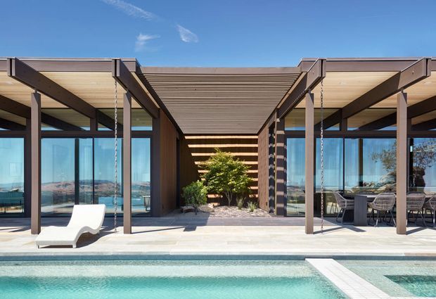 A modern single-storey pavilion-style residence arranged around a covered outdoor passage and swimming pool. Deep timber-lined pergolas and dark structural posts frame extensive floor-to-ceiling glazing, with an outdoor dining area on the right and a landscaped courtyard visible centrally. The pool terrace overlooks a bright blue waterfront or coastal landscape in the distance.