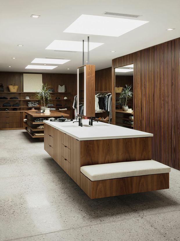A refined walk-in wardrobe or dressing room centered on a large walnut-toned island with white benchtop, integrated drawers, a cushioned seating ledge, and a mirrored central display cabinet. Dark timber shelving and cabinetry line the rear wall, while a vertical timber slat feature wall and full-height mirror add texture and depth. Clothing rails, accessories, indoor plants, recessed lighting, and large rectangular skylights are visible. The floor is a pale polished concrete or terrazzo-like finish, creating a warm contemporary interior.