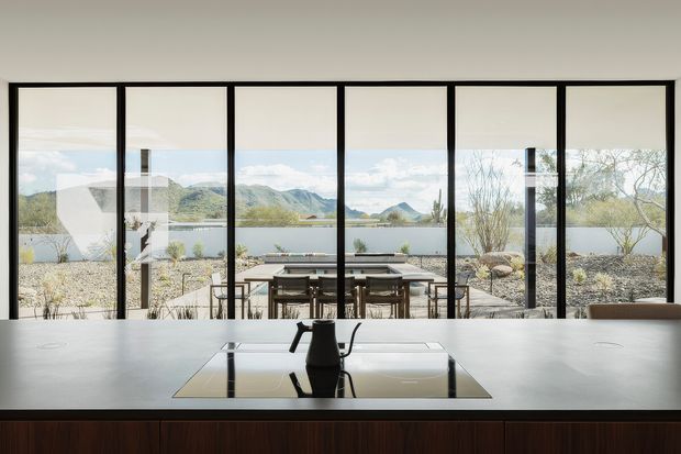 A refined modern interior viewed from behind a large work or dining table, with a black office chair in the foreground and full-height black-framed glazing opening to a timber outdoor dining deck. A lake, mountainous landscape, and sparse landscaped planting are visible beyond the glazing. The room is bright and minimalist, with dark timber fixed joinery beneath the broad pale tabletop and strong horizontal and vertical window framing.