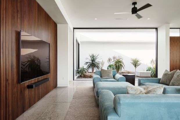 Bright contemporary living room with a large pale blue sectional sofa and matching armchairs arranged around a television. The TV wall features vertical dark timber slat panelling and a floating media shelf. Full-height glazing opens toward a planted courtyard, while a ceiling fan, pale flooring, neutral walls, and soft furnishings create an airy, relaxed interior.