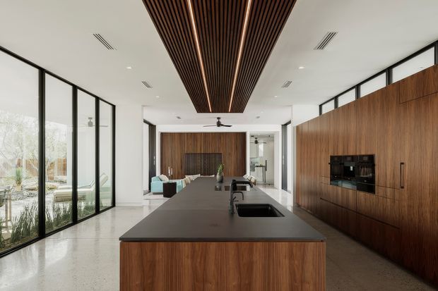 A spacious contemporary open-plan kitchen and living area with a large dark stone island, integrated sink and tap, and extensive warm timber cabinetry. Full-height black-framed glazing lines the left side, while a dramatic slatted timber ceiling feature extends above the island. The polished light floor, white ceiling, black-framed openings, and wood wall panelling create a refined indoor-outdoor atmosphere, with a lounge area visible beyond the kitchen.