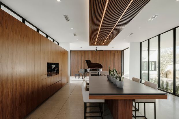 A refined open-plan interior combining a dining area, kitchen joinery, and a piano zone. Full-height warm timber cabinetry lines the left wall and continues across the rear, while a long dark stone dining table with timber detailing occupies the foreground. A black grand piano sits at the back, and large floor-to-ceiling windows bring in abundant natural light. A dramatic linear timber slatted ceiling feature adds warmth and architectural definition to the otherwise clean, contemporary space.