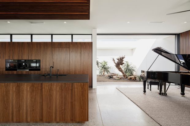 A refined open-plan interior featuring a warm timber kitchen with full-height wood-grain cabinetry, dark stone benchtops, integrated appliances, and a central island. The kitchen opens into a bright living area with a black grand piano, pale flooring, and expansive glazing overlooking a landscaped courtyard. A slatted timber ceiling feature adds architectural warmth and visual interest.