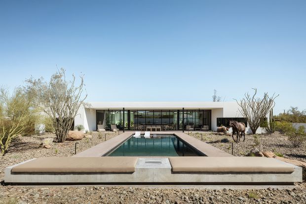Single-storey modern residence set in an arid desert landscape, viewed straight on across a long rectangular reflecting pool and raised concrete seating platform. The building has a low, flat roof, white rendered walls, extensive dark-framed floor-to-ceiling glazing, and a symmetrical linear composition. Sparse desert planting, gravel, sculptural cacti, and dry-climate trees surround the house. The pool and strong axial layout create a calm, minimalist architectural scene under a clear blue sky.