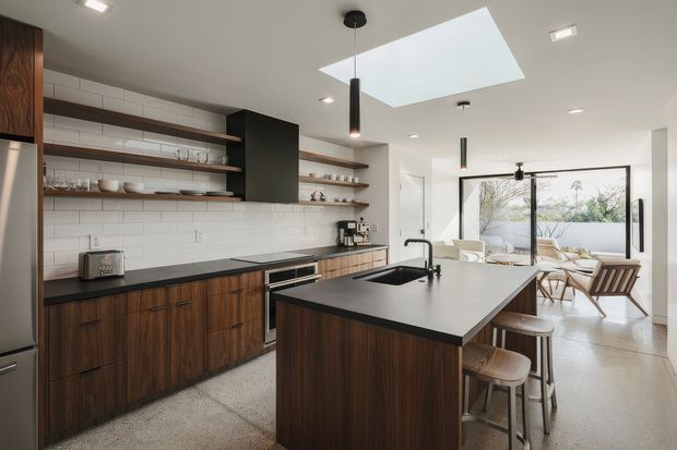 A bright contemporary kitchen with dark timber cabinetry, a large black island with integrated sink and seating, white subway-tile splashback, open timber shelves, and terrazzo-style flooring. A large skylight and broad glazed opening connect the kitchen to a light-filled dining and outdoor area.
