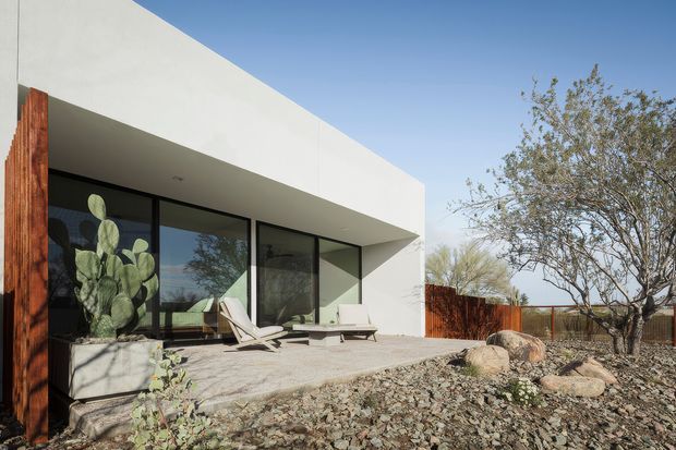 Exterior view of a minimalist, flat-roofed modern residence with a crisp white rendered facade, deep roof overhang, and expansive dark-framed glazing opening onto a paved terrace. The terrace has outdoor lounge chairs and a low built-in concrete planter, while a weathered timber privacy fence and sparse xeric landscaping with rocks and mature trees surround the home.