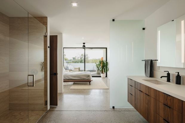 Contemporary ensuite bathroom viewed from the shower area, with a frameless glass shower enclosure, large-format beige stone-look wall tiles, concrete-look flooring, and a floating dark timber double vanity with white benchtop and black tapware. A bedroom is clearly visible directly beyond the open bathroom, separated by a timber door and glass partition. The space is bright, refined, and minimalist with natural materials and strong indoor-outdoor glazing.