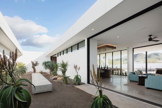 A bright contemporary courtyard and open-plan living area with strong indoor–outdoor connection. White minimalist buildings frame a landscaped gravel courtyard with sculptural agave and succulent planting, a long concrete bench, and expansive black-framed glazing. Through the open sliding doors, a refined living and dining space is visible, with pale flooring, a dark kitchen island or bar, turquoise lounge chairs, and views across water to mountainous hills.