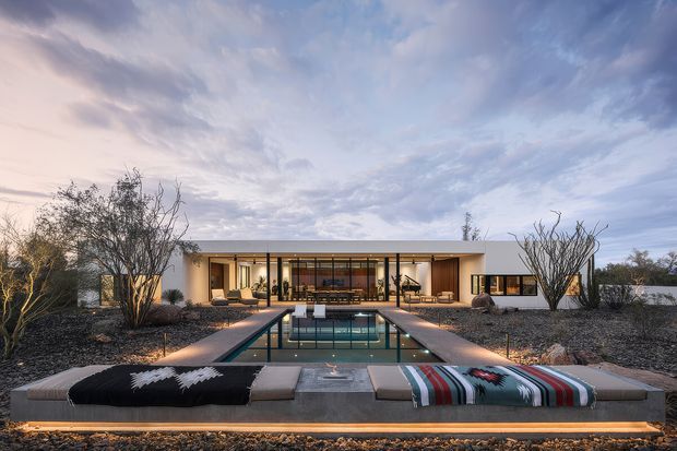 A low, single-storey modern residence set in an arid landscaped site, viewed from across a long rectangular swimming pool. The house has a horizontal flat-roofed form, expansive glazed central living area, pale rendered walls, dark-framed openings, and warm interior lighting. A raised concrete poolside platform in the foreground holds upholstered outdoor loungers with striped and patterned throws, while sparse sculptural desert planting and gravel surround the building.