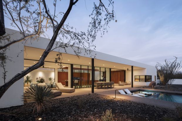 A modern single-storey residence viewed from the landscaped exterior at dusk, with a broad flat roof, white rendered walls, extensive floor-to-ceiling glazing, and warm timber interior joinery. The covered outdoor living area opens onto a patio with lounge seating, dining furniture, and a rectangular swimming pool. Gravel landscaping, sculptural drought-tolerant planting, and a mature tree frame the composition.