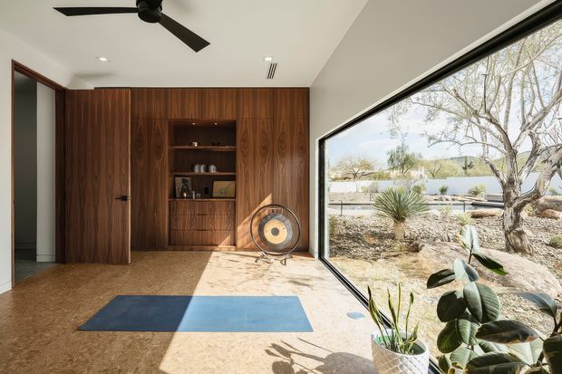 A bright, minimalist interior featuring a richly grained dark timber built-in wall with concealed storage, a central open display niche and ribbed cabinet doors. A large corner window provides expansive views of a dry landscaped garden and distant hills. The room has a polished concrete floor, white ceiling with recessed downlights and a black ceiling fan, plus a blue exercise mat and indoor plants. The space appears to function as a home gym or flexible wellness room.
