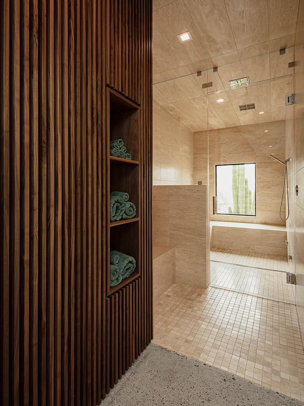 A refined walk-in shower bathroom viewed from an adjoining space. The shower enclosure is fully lined in warm beige stone-look tiles, with matching small-format mosaic floor tiles, a frameless glass screen and recessed ceiling lighting. A dark timber vertical-slat wall with open shelving and neatly folded towels forms a strong feature element at the entrance. A terrazzo-like grey floor is visible outside the shower.