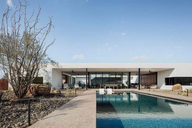 A low, minimalist modern residence surrounding a long rectangular swimming pool. The flat-roofed pavilion has extensive floor-to-ceiling glazing, dark window frames, a sheltered outdoor living and dining area, and pale rendered walls. The foreground includes a broad concrete pool terrace and xeriscaped landscaping with gravel, boulders, and sparse sculptural trees.