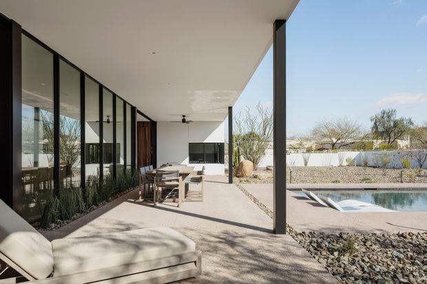 A covered outdoor terrace adjoining a modern glass-walled residence, with a dining table, built-in outdoor fireplace or media wall, lounge seating, and a swimming pool beyond. The space has a flat roof, slender dark structural columns, expansive floor-to-ceiling glazing, and a dry landscaped setting with gravel and drought-tolerant planting.