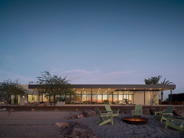 A single-storey modern residence viewed from an expansive landscaped outdoor area at dusk. The building has a long, low flat roof, extensive floor-to-ceiling glazing, warm interior lighting, and a restrained neutral facade. In the foreground are gravel and sandy outdoor zones with boulders, casual green Adirondack-style chairs, and a circular fire bowl. Sparse planting and sculptural palms contribute to a desert-like setting.
